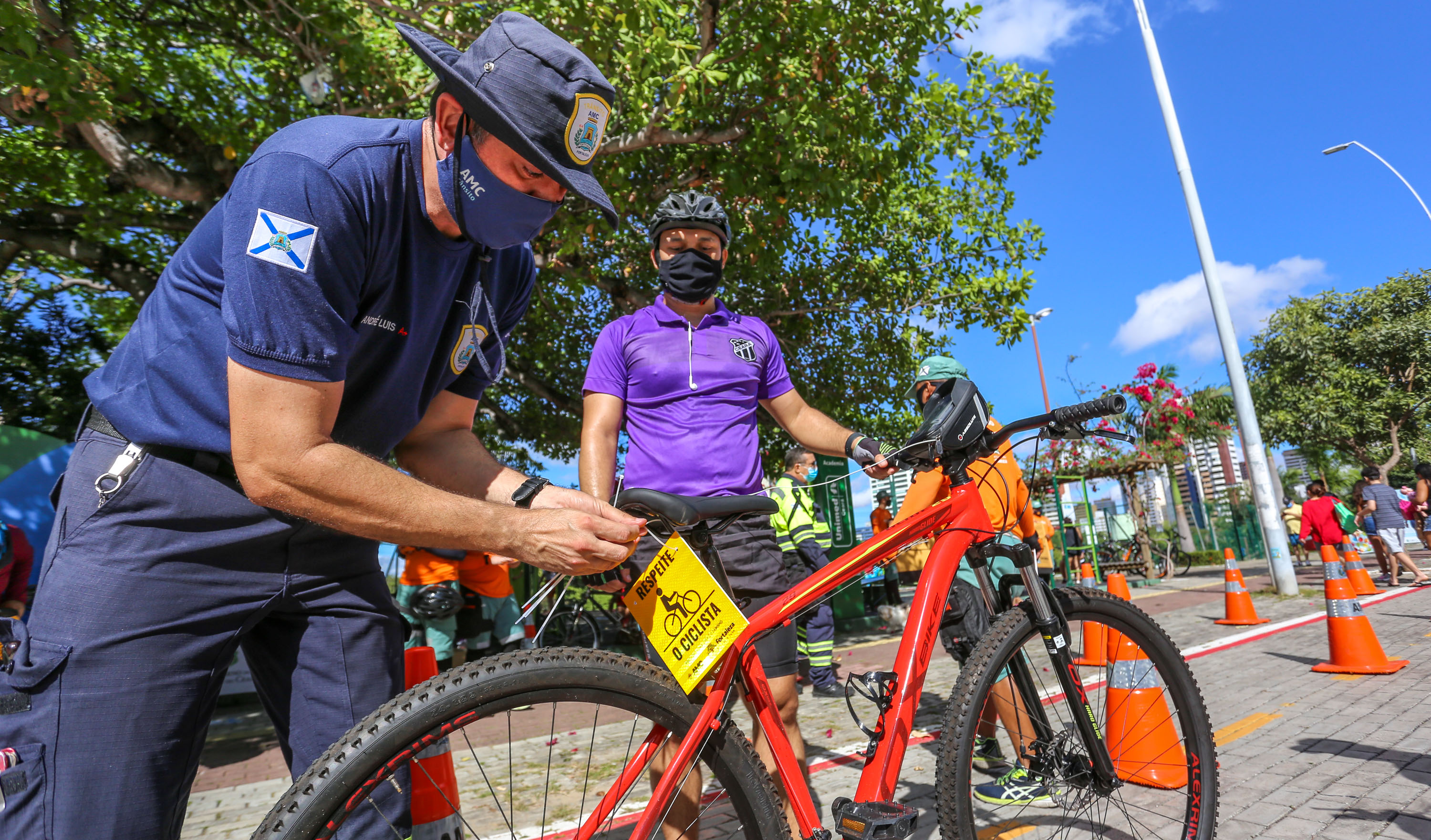 agente da AMC coloca uma plaquinha atrás do selim de uma bicicleta enquanto é observado pelo ciclista